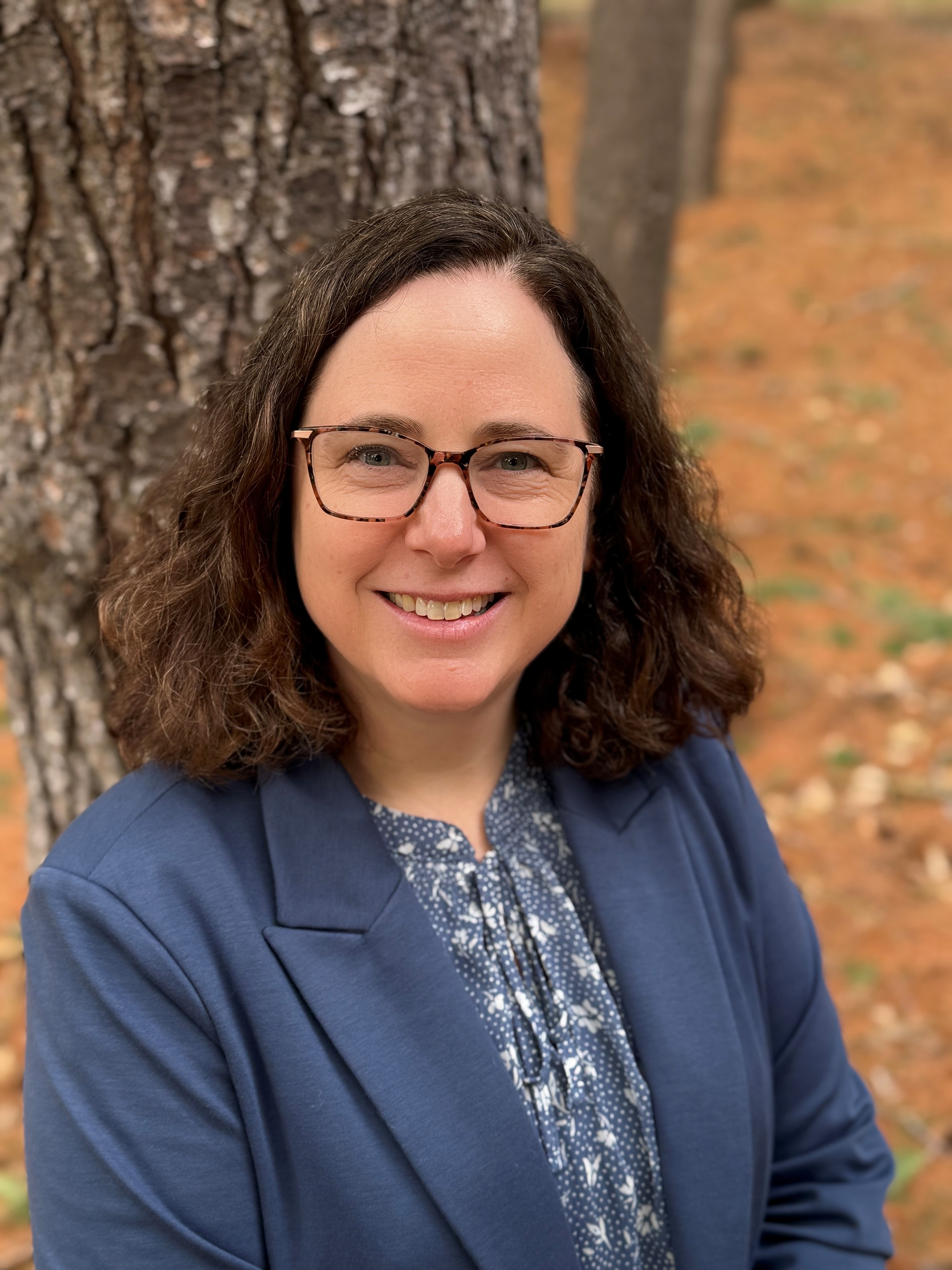 Outdoor portrait of Rose Stewart standing near a tree with fallen leaves on the ground in the background.